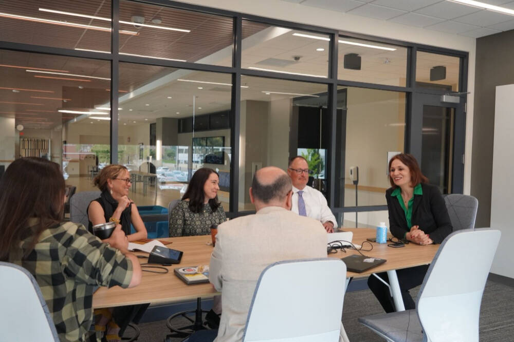 A group of six professionals sit around a conference table engaged in a discussion inside a modern office space with large windows and glass walls. The group includes both men and women, and they appear to be collaborating in a meeting.